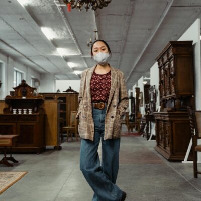 A woman wearing a mask stands inside an antique store surrounded by vintage wooden furniture.
