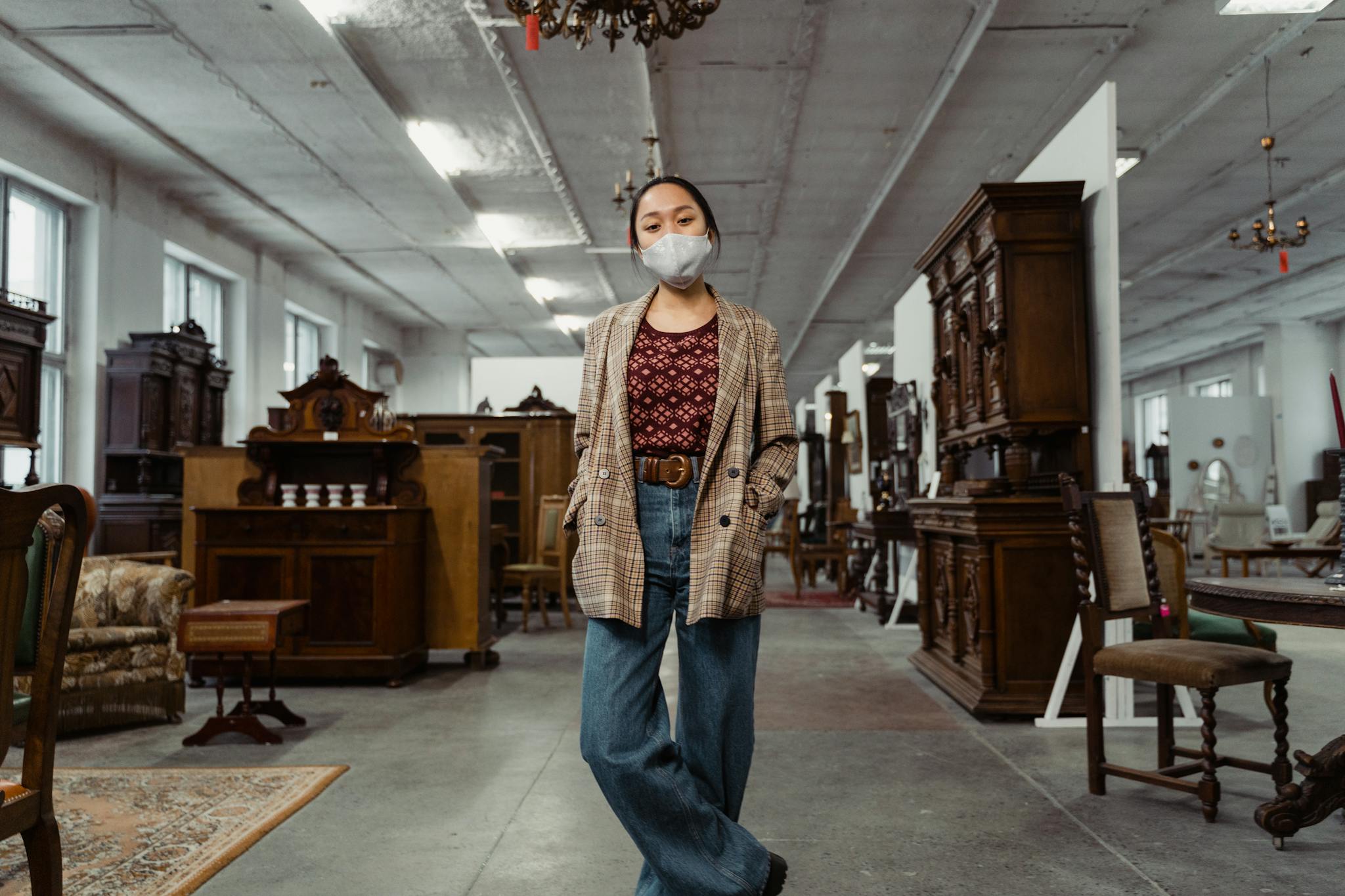A woman wearing a mask stands inside an antique store surrounded by vintage wooden furniture.